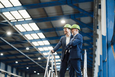 Smiling male business professionals wearing hardhat standing by railing in factory - JOSEF05721