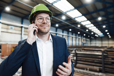 Lächelnder Geschäftsmann, der in einer Fabrik mit seinem Smartphone telefoniert - JOSEF05725
