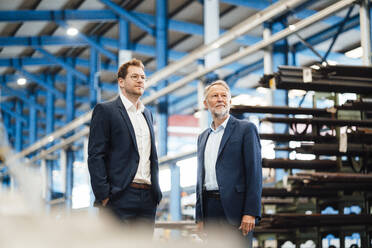 Mid adult businessman standing with hands in pockets by male colleague in factory - JOSEF05731