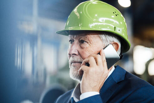 Männlicher Manager mit Schutzhelm, der in einer Fabrik mit einem Mobiltelefon spricht - JOSEF05758