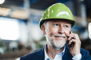 Lächelnder männlicher Manager, der in einer Fabrik mit einem Smartphone telefoniert - JOSEF05759