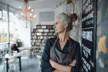 Female cafe owner with arms crossed leaning at blackboard in coffee shop - JOSEF05798