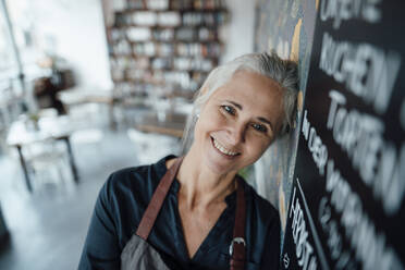 Smiling female cafe owner wearing apron leaning on blackboard in coffee shop - JOSEF05799