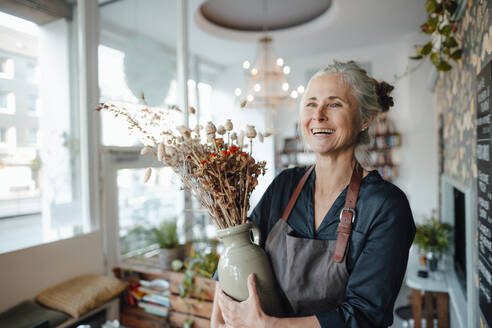 Smiling female cafe owner holding flower vase in coffee shop - JOSEF05807