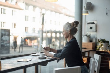 Eine Freiberuflerin benutzt ein Mobiltelefon, während sie in einem Café am Glasfenster sitzt - JOSEF05880