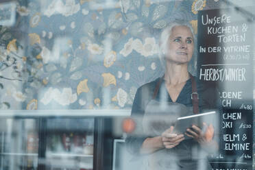 Weibliche Café-Besitzerin hält digitales Tablet, während sie in einem Café steht, gesehen durch ein Glasfenster - JOSEF05888