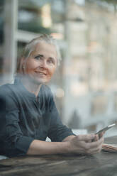 Freiberuflerin mit Mobiltelefon im Café sitzend durch Glasfenster gesehen - JOSEF05898