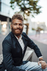 Smiling mature businessman with brown hair sitting on bench - GUSF06374