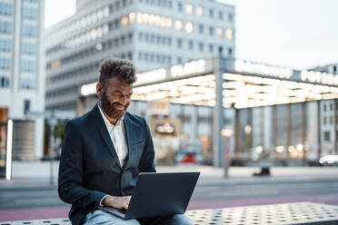 Lächelnder Geschäftsmann arbeitet am Laptop am Bahnhof - GUSF06378