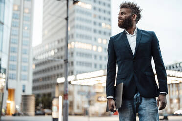 Male freelancer with laptop standing in front of building - GUSF06381