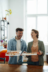 Cheerful man and woman holding pizza by table at home - GUSF06427