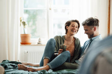 Happy woman with mug sitting with man on bed at home - GUSF06435