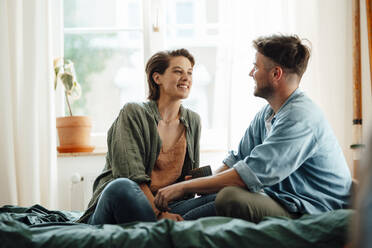 Happy mid adult couple talking while sitting on bed at home - GUSF06436