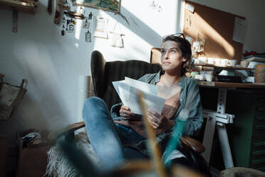Weibliche Fachkraft sitzt auf einem Stuhl und hält eine Skizze zu Hause im Büro - GUSF06456