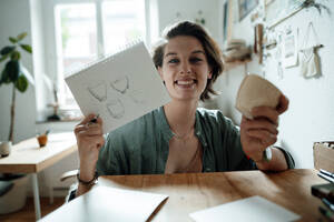 Smiling female illustrator holding spiral notebook and cup on table at home office - GUSF06462