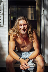 Shirtless young man smiling while sitting at doorway of motor home - PGF00783