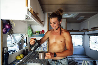 Lächelnder Mann ohne Hemd gießt Kaffee ein, während er im Wohnmobil steht - PGF00788