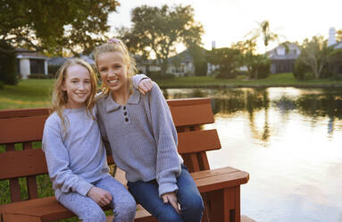 Smiling girls sitting on bench by pond - AZF00360