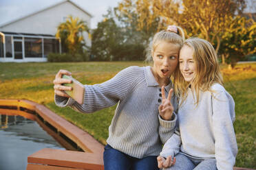 Mädchen mit offenem Mund gestikuliert bei der Aufnahme von Selfie mit Freund durch Smartphone durch Teich - AZF00362
