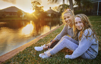 Happy girls sitting on grass by pond during sunset - AZF00363