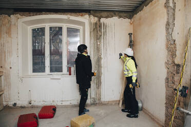 Weibliche Bauunternehmerin diskutiert mit einem männlichen Arbeiter, während sie auf der Baustelle eine Wand untersucht - MASF25132