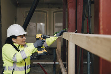 Female worker using drill on wooden plank while working at construction site - MASF25138