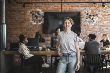 Female entrepreneur standing with arms crossed while colleagues working in creative office - MASF25214