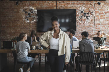 Female entrepreneur standing with arms crossed while colleagues working in creative office - MASF25216