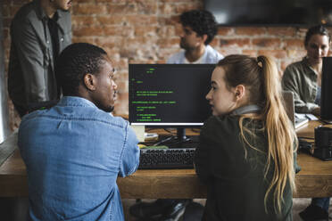 Junger weiblicher Hacker im Gespräch mit einem männlichen Kollegen beim Programmieren am Computer im Büro - MASF25249
