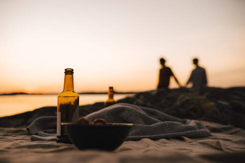 Bierflasche und Schüssel auf Picknickdecke, während schwules Paar im Hintergrund am Seeufer bei Sonnenuntergang - MASF25322