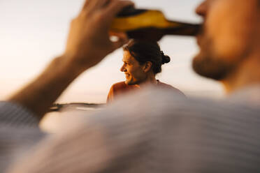 Man drinking beer while smiling male friend looking away at lakeshore in evening - MASF25332