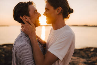 Lächelndes homosexuelles Paar, das am Seeufer bei Sonnenuntergang von Angesicht zu Angesicht steht - MASF25337
