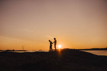 Male friends holding hands while walking at lakeshore during sunset - MASF25339
