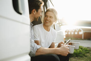 Lächelndes schwules Paar mit Kaffeetasse verbringt seine Freizeit im Campingwagen an einem sonnigen Tag - MASF25368