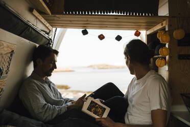 Mann sitzt mit Buch, während sein Freund im Wohnmobil sein Smartphone benutzt - MASF25377