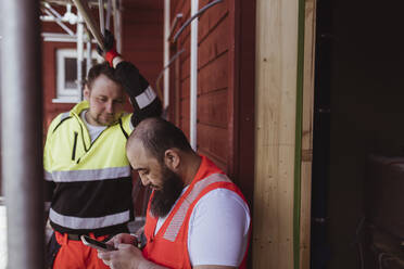 Männlicher Bauarbeiter schaut auf einen bärtigen Kollegen, der auf der Baustelle ein Smartphone benutzt - MASF25390