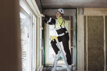Bauunternehmerin in voller Länge bei der Vermessung von Fensterrahmen auf einer Stufenleiter auf der Baustelle - MASF25399