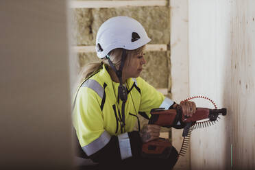 Female builder using work tool on wall while working at construction site - MASF25415