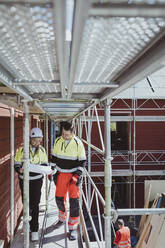 Full length of female building contractor discussing over floor plan with male colleague while walking at scaffolding - MASF25421