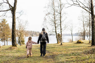 Full length rear view of mother and daughter holding hands while walking towards lake - MASF25532