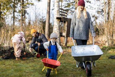 Smiling woman and son wheeling wheelbarrows while family gardening in background at back yard - MASF25536