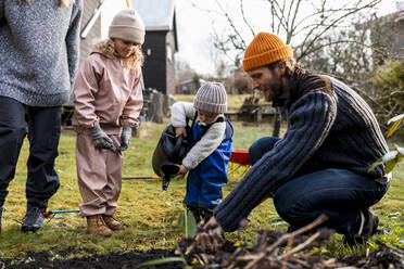 Mann führt Sohn mit Gießkanne durch Mädchen bei der Gartenarbeit im Hinterhof - MASF25539