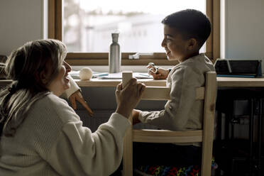 Lächelnder Sohn im Gespräch mit seiner Mutter, während er zu Hause auf einem Stuhl sitzt - MASF25575