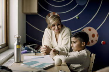 Lächelnde Mutter schaut auf ihren autistischen Sohn, der zu Hause am Tisch sitzt - MASF25580