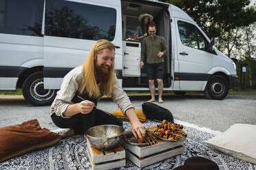 Bärtiger Mann bereitet Fleischspieße zum Grillen vor, während Freunde im Wohnmobil sitzen - MASF25653