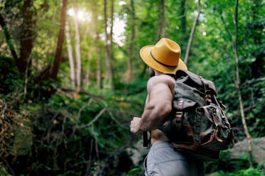 Von unten von einem fitten männlichen Reisenden ohne Hemd mit Rucksack und Hut, der in einem felsigen Wald steht und wegschaut - ADSF29954