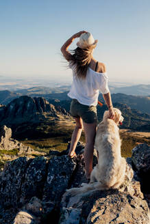 Back view of young woman with Golden Retriever dog standing on rock and admiring breathtaking mountains during summer trip - ADSF29956