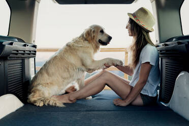 Barefoot woman holding paw of loyal Golden Retriever dog while sitting on bed inside RV during road trip in nature - ADSF29959