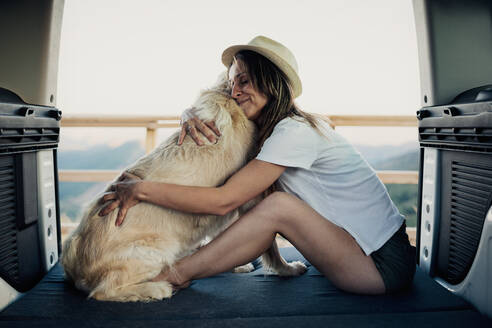 Barefoot woman embracing loyal Golden Retriever dog while sitting on bed inside RV during road trip in nature - ADSF29960