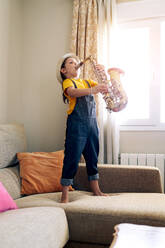 Seitenansicht eines barfüßigen Kindes, das Saxophon spielt, während es zu Hause auf der Couch steht (tagsüber) - ADSF29962